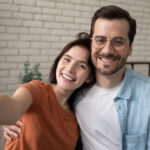Young happy couple with big smiles. Taking a selfie in front of a white brick wall.