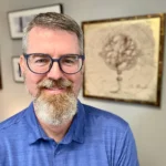 Professional man wearing a blue polo shirt and glasses. He is smiling and standing in front of art hung on the wall.